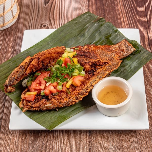 Whole fried fish on a banana leaf, topped with diced tomatoes, onions, and green onions, with a side of sauce.