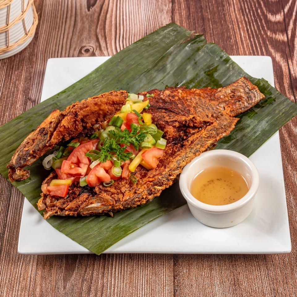 Whole fried fish on a banana leaf, topped with diced tomatoes, onions, and green onions, with a side of sauce.
