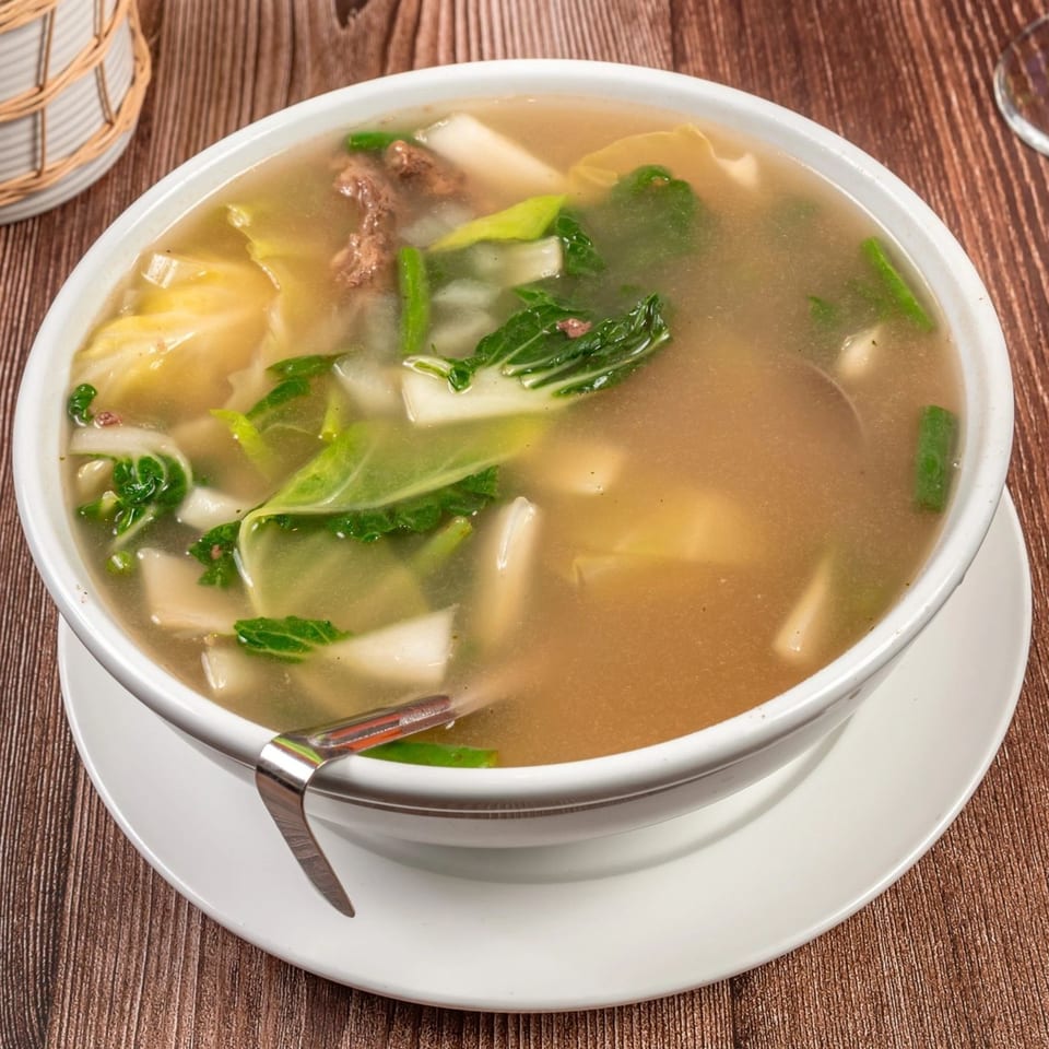 A white bowl of Filipino nilaga soup with beef, cabbage, and bok choy on a wooden table.