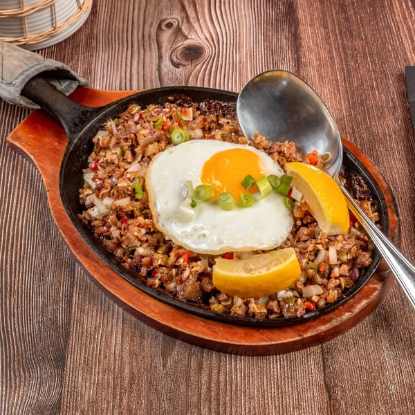 Filipino sizzling sisig with a fried egg, lemon wedges, and a serving spoon on a wooden table.