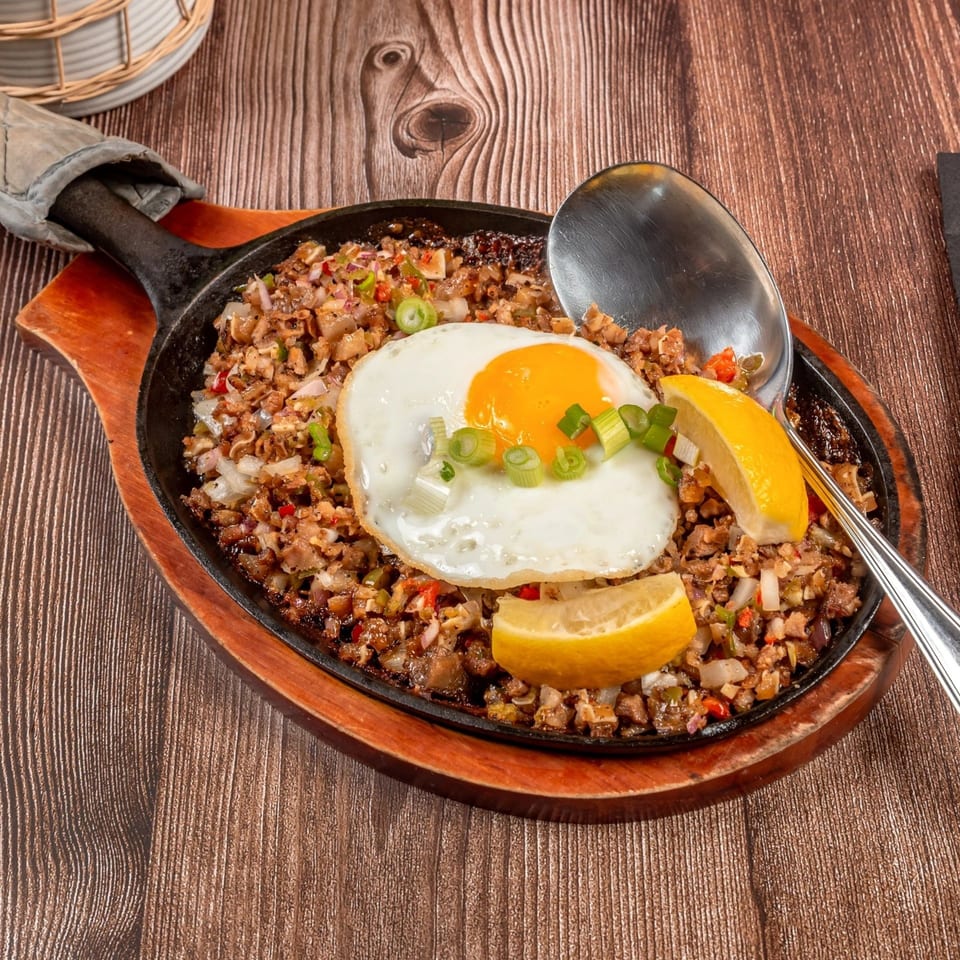 Filipino sizzling sisig with a fried egg, lemon wedges, and a serving spoon on a wooden table.
